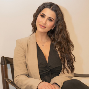 Dr Amani with dark brown hair, wearing a beige blazer over a black top, sits in a dark wooden chair.