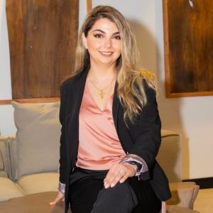 Dr Nora in a black blazer and a blush-pink top sits on a light beige couch, smiling confidently.