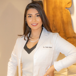 Smiling female dentist, Dr. Yalda Saberi, in a white lab coat, stands against a wooden background.