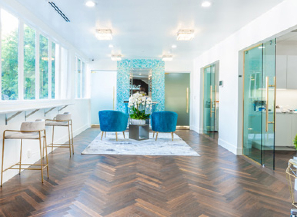 Modern, light-filled interior with teal accent chairs, a herringbone wood floor, and glass partitions.