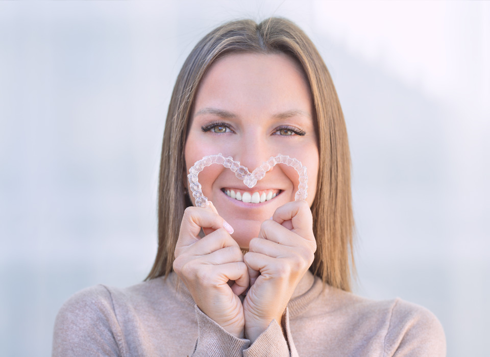 Woman with a happy expression holds a clear heart-shaped dental aligner in front of her face.