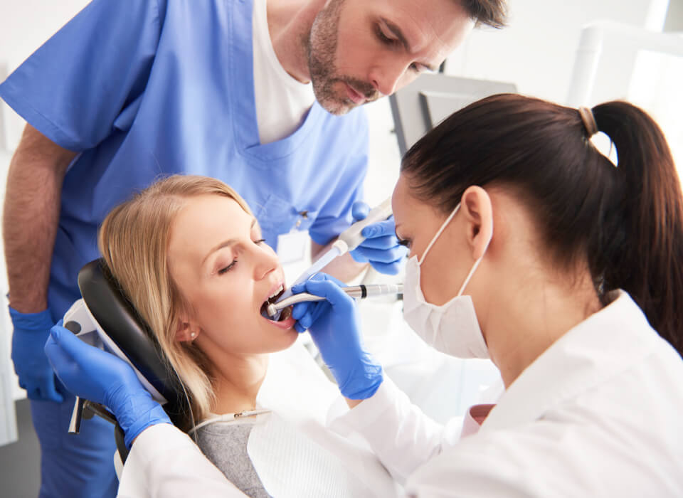 Dental procedure in progress; woman patient with open mouth, dentists in blue and white scrubs using dental tools.