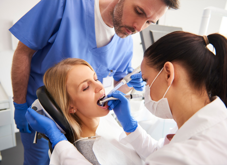 Dental procedure in progress; woman patient with open mouth, dentists in blue and white scrubs using dental tools.