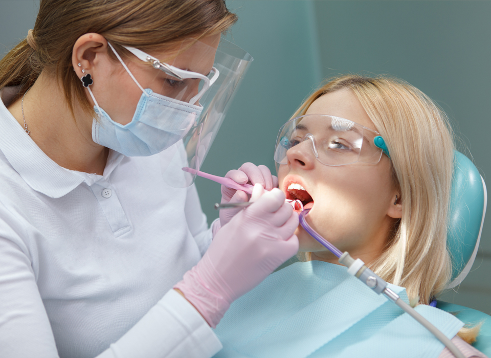 Dental hygienist performing oral exam on a patient wearing protective eyewear and a mask.