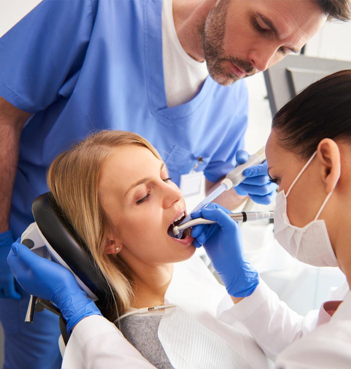 Dental procedure in progress; woman patient with open mouth, dentists in blue and white scrubs using dental tools.